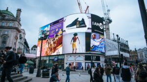 a bustling urban scene at a major public square, highlighted by a large, multi-panel digital billboard displaying various advertisements. People of diverse ages and attire mill about the square, some engaged with their phones, others conversing or simply passing through.