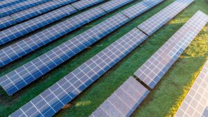 An aerial view of a large solar farm at sunset, showcasing long rows of photovoltaic panels installed on a grassy field, with the golden hues of the sunlight casting a warm glow over the scene.